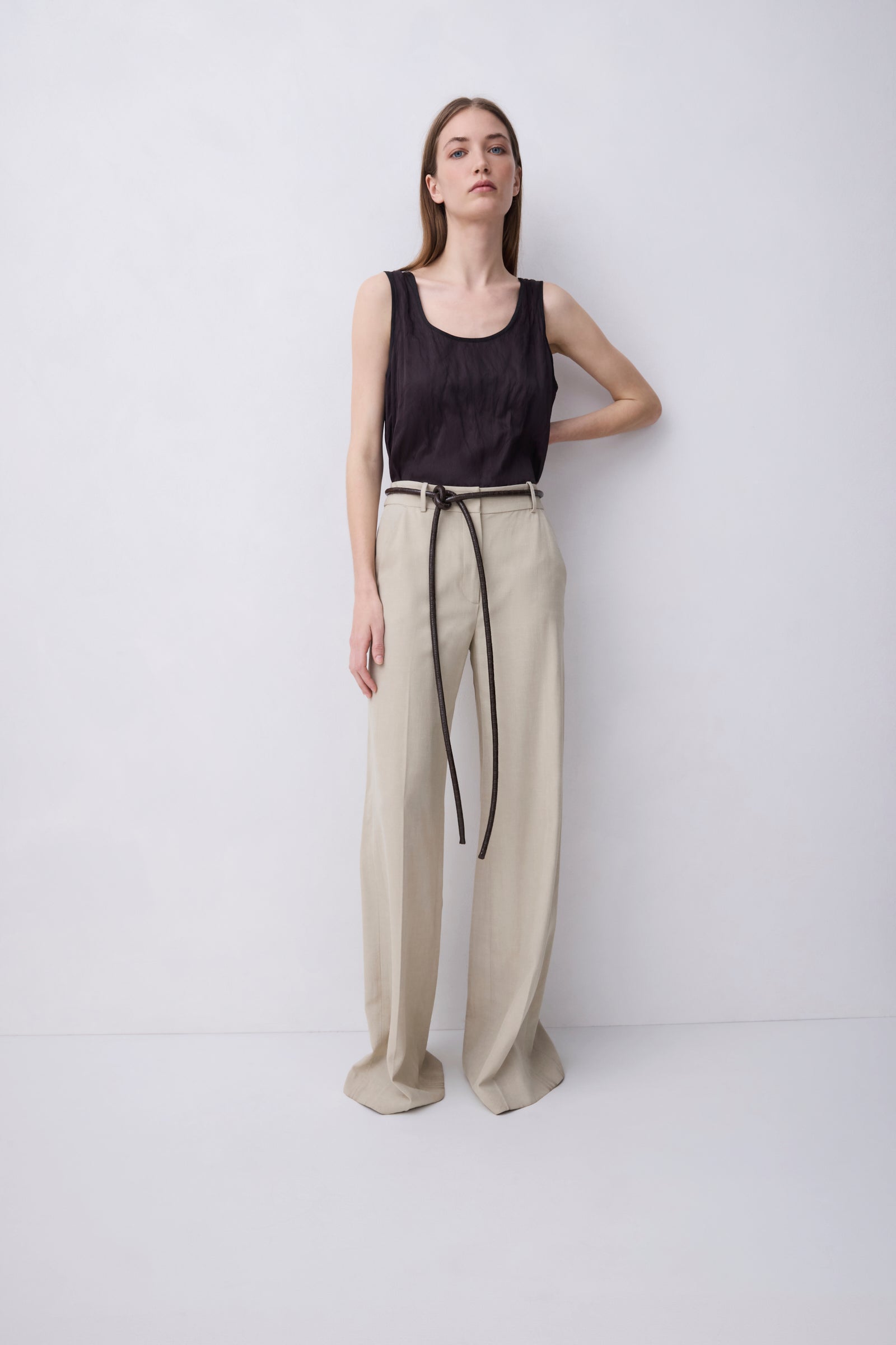 A woman stands against a plain light gray studio background wearing a sleeveless black top and wide-leg beige trousers with a thin dark tie belt.