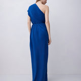 A woman modeling a floor-length, one-shoulder blue gown from the back against a plain light gray studio background.