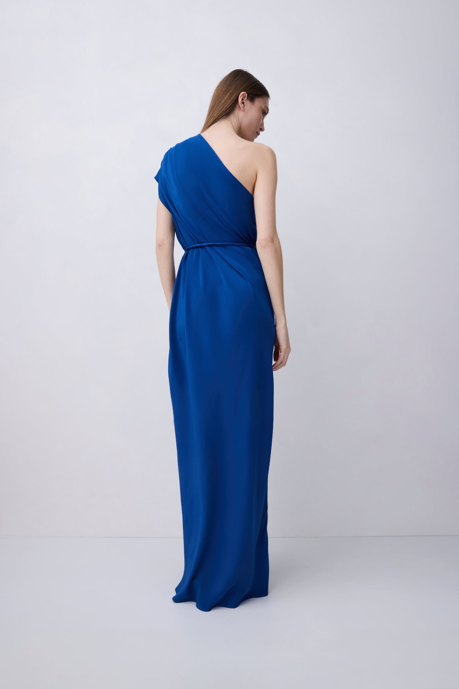 A woman modeling a floor-length, one-shoulder blue gown from the back against a plain light gray studio background.