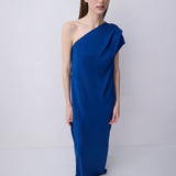 A woman modeling a floor-length, one-shoulder draped dress in a rich royal blue color, set against a plain light gray studio background.