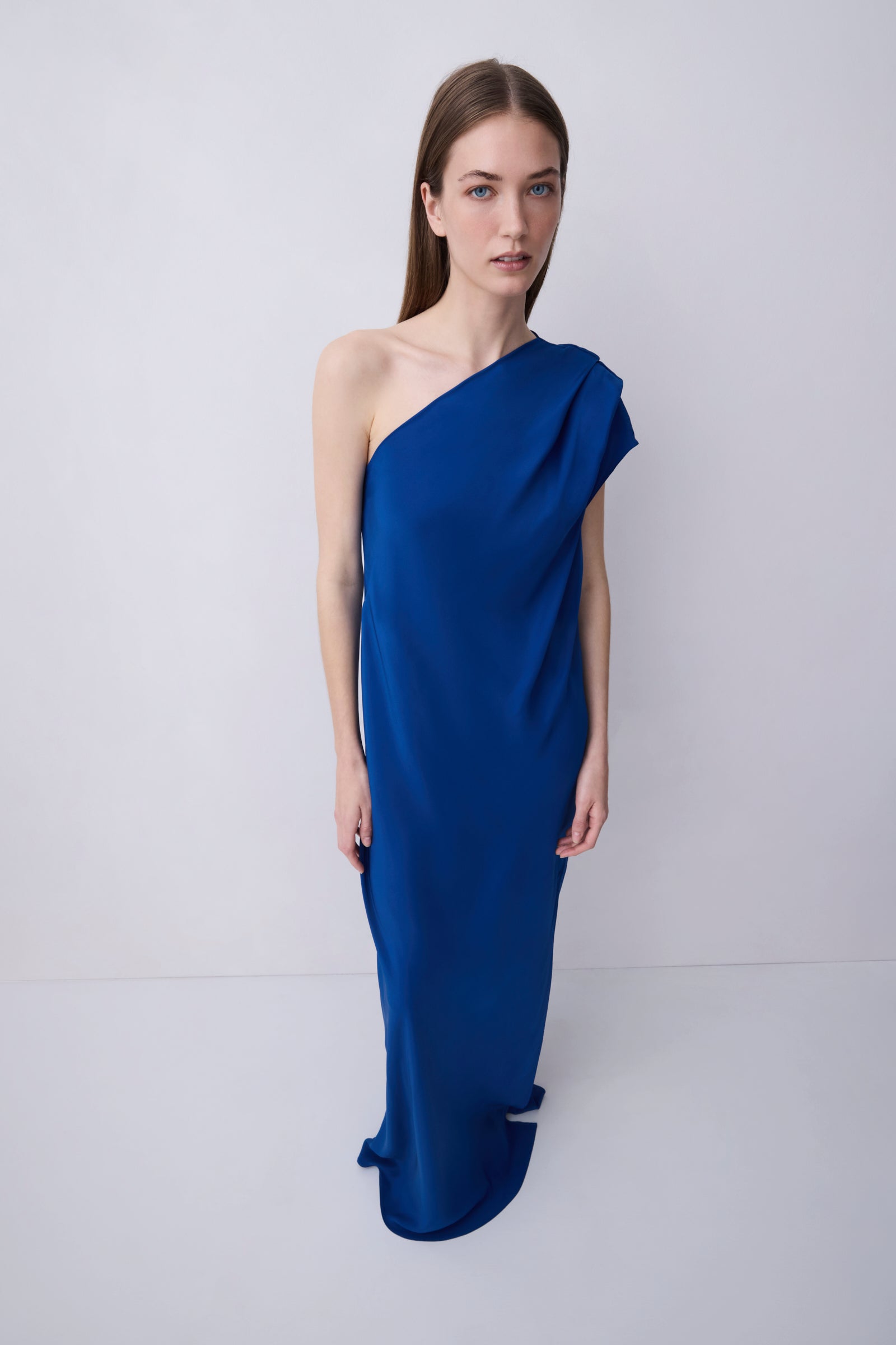 A woman modeling a floor-length, one-shoulder draped dress in a rich royal blue color, set against a plain light gray studio background.