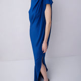 A woman modeling a draped, one-shoulder midi dress in a vibrant royal blue, paired with black strappy flat sandals, set against a plain light gray studio background.