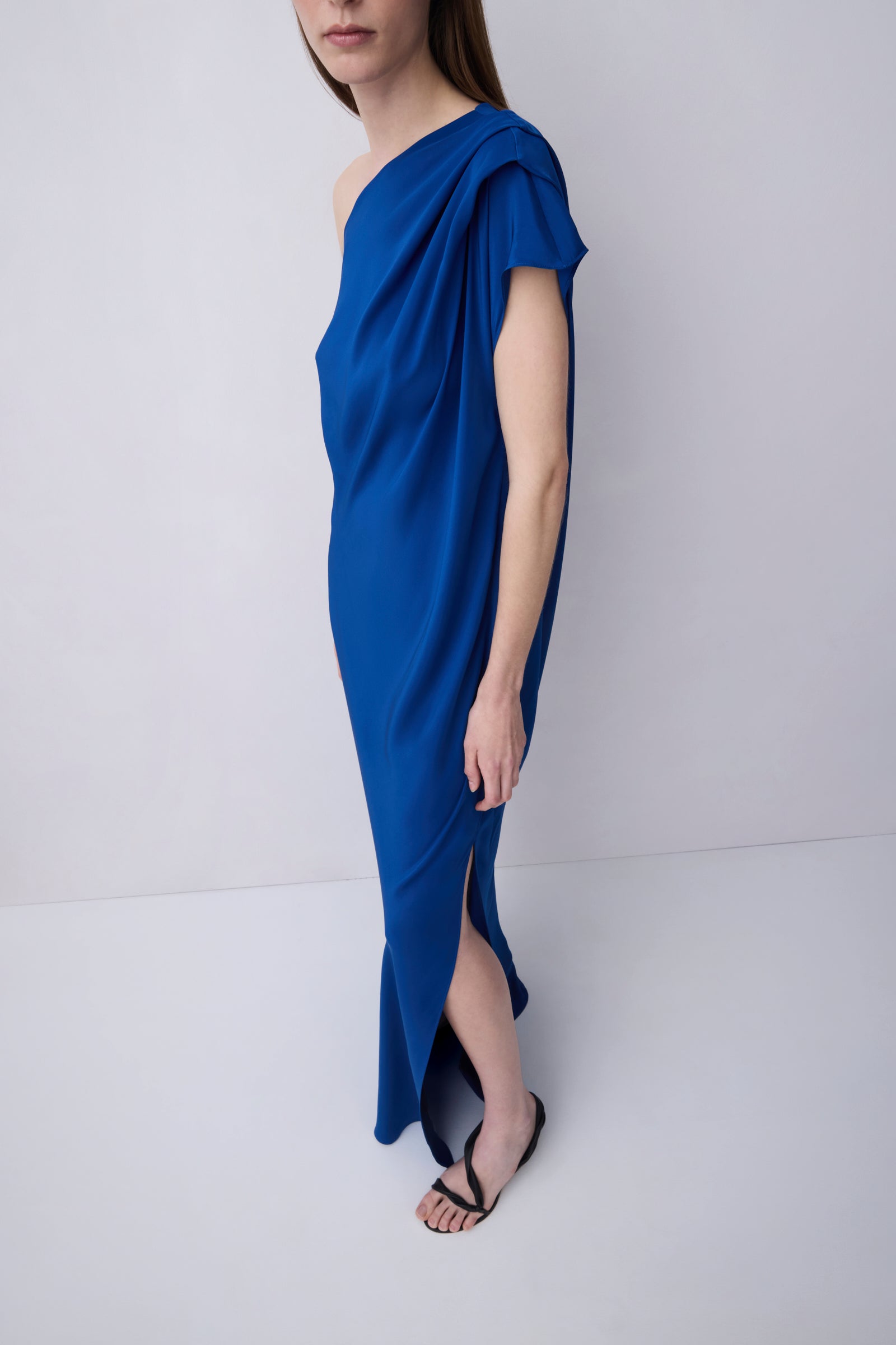 A woman modeling a draped, one-shoulder midi dress in a vibrant royal blue, paired with black strappy flat sandals, set against a plain light gray studio background.