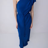 A woman modeling a royal blue one-shoulder midi dress with a draped neckline, a matching tie belt at the waist, and a flowing skirt, set against a plain light gray studio background.