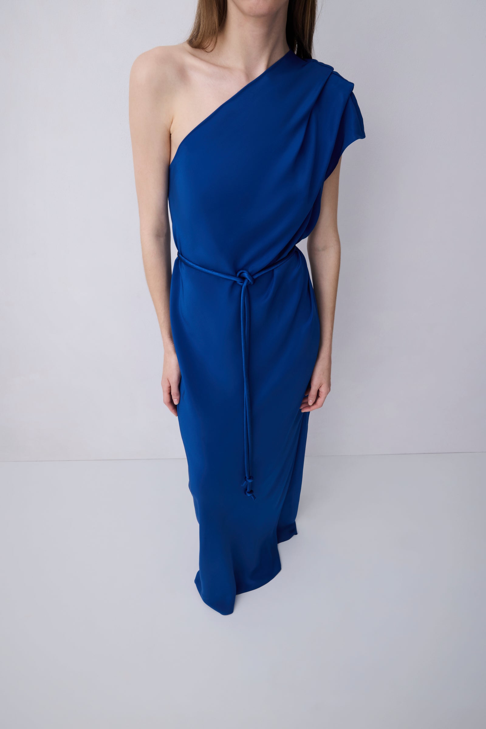A woman modeling a royal blue one-shoulder midi dress with a draped neckline, a matching tie belt at the waist, and a flowing skirt, set against a plain light gray studio background.