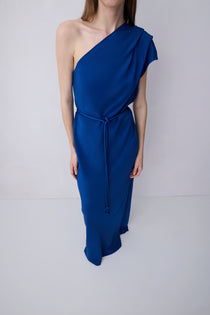 A woman modeling a royal blue one-shoulder midi dress with a draped neckline, a matching tie belt at the waist, and a flowing skirt, set against a plain light gray studio background.