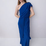 A woman modeling a royal blue one-shoulder maxi dress with a draped neckline, short cap sleeves, a matching waist tie, and a flowing skirt, set against a plain light gray studio background.