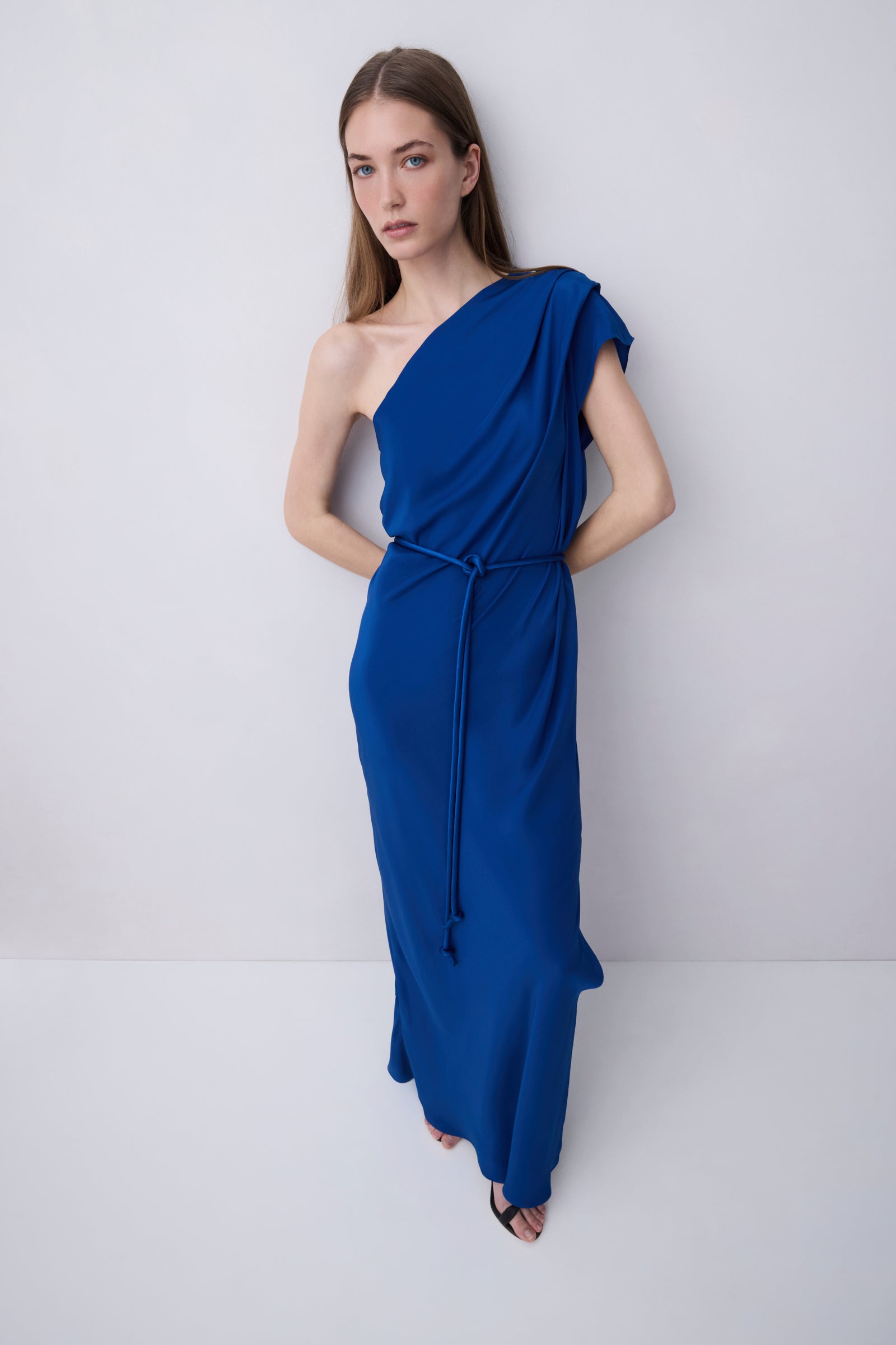 A woman modeling a royal blue one-shoulder maxi dress with a draped neckline, short cap sleeves, a matching waist tie, and a flowing skirt, set against a plain light gray studio background.