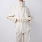 A model wearing a cream-colored high-collar jacket with voluminous sleeves and matching wide-leg trousers, set against a plain light gray studio background.