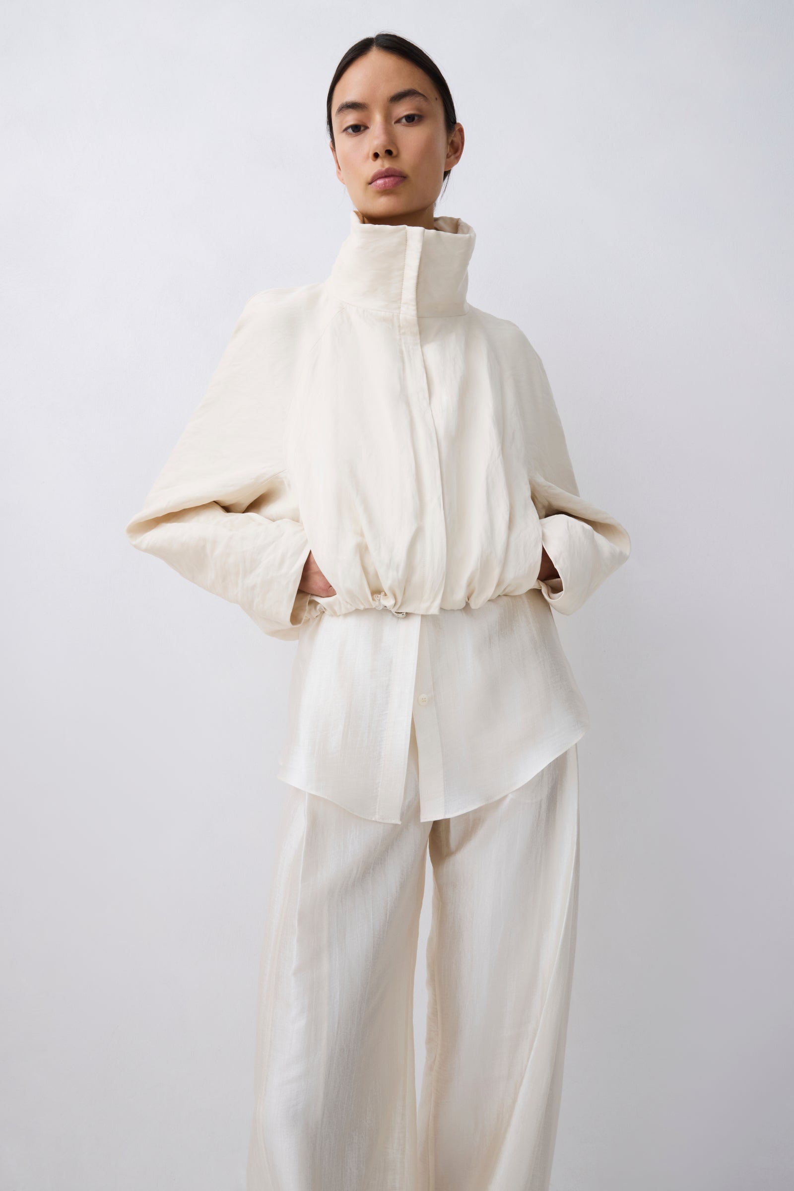 A model wearing a cream-colored high-collar jacket with voluminous sleeves and matching wide-leg trousers, set against a plain light gray studio background.