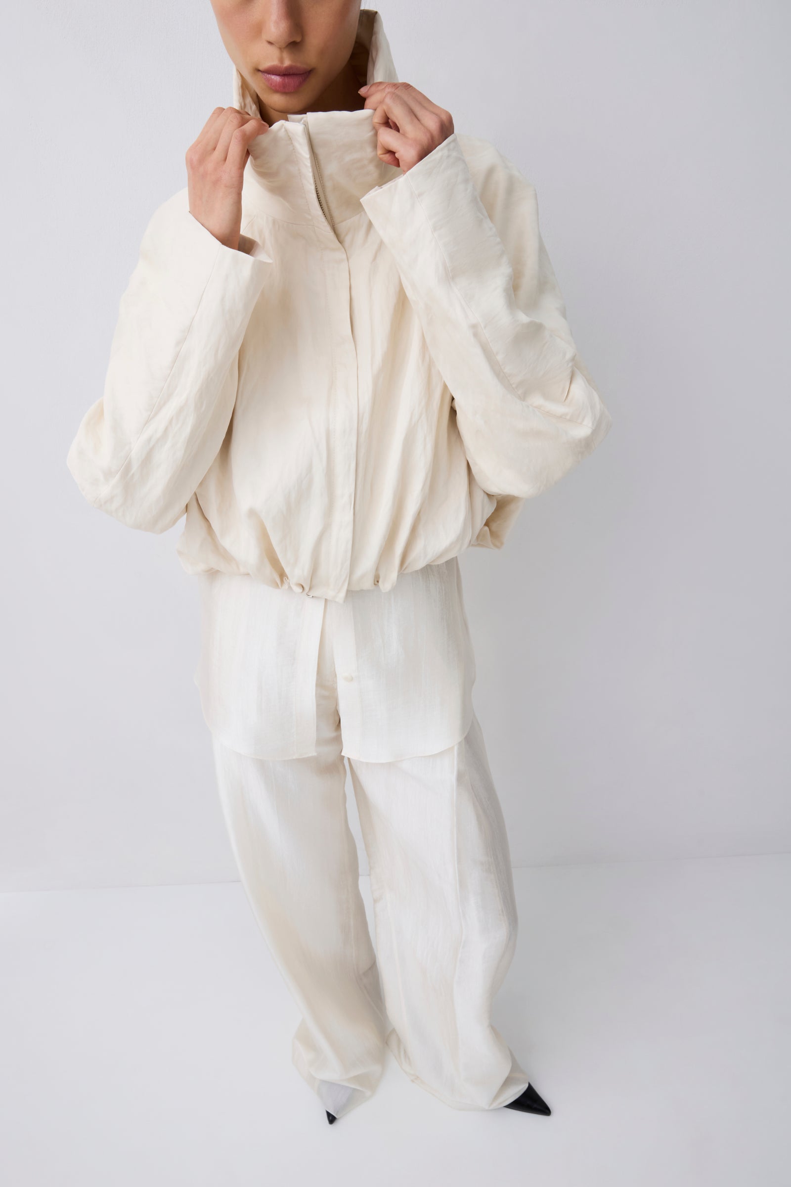 A model wearing a cream-colored, textured two-piece outfit consisting of a cropped zip-up jacket and matching wide-leg trousers, photographed against a neutral light gray background.