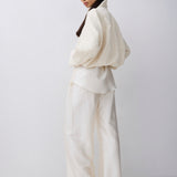 A model wearing a cream-colored two-piece outfit consisting of a voluminous long-sleeve top and matching wide-leg trousers, photographed against a plain light gray studio background.