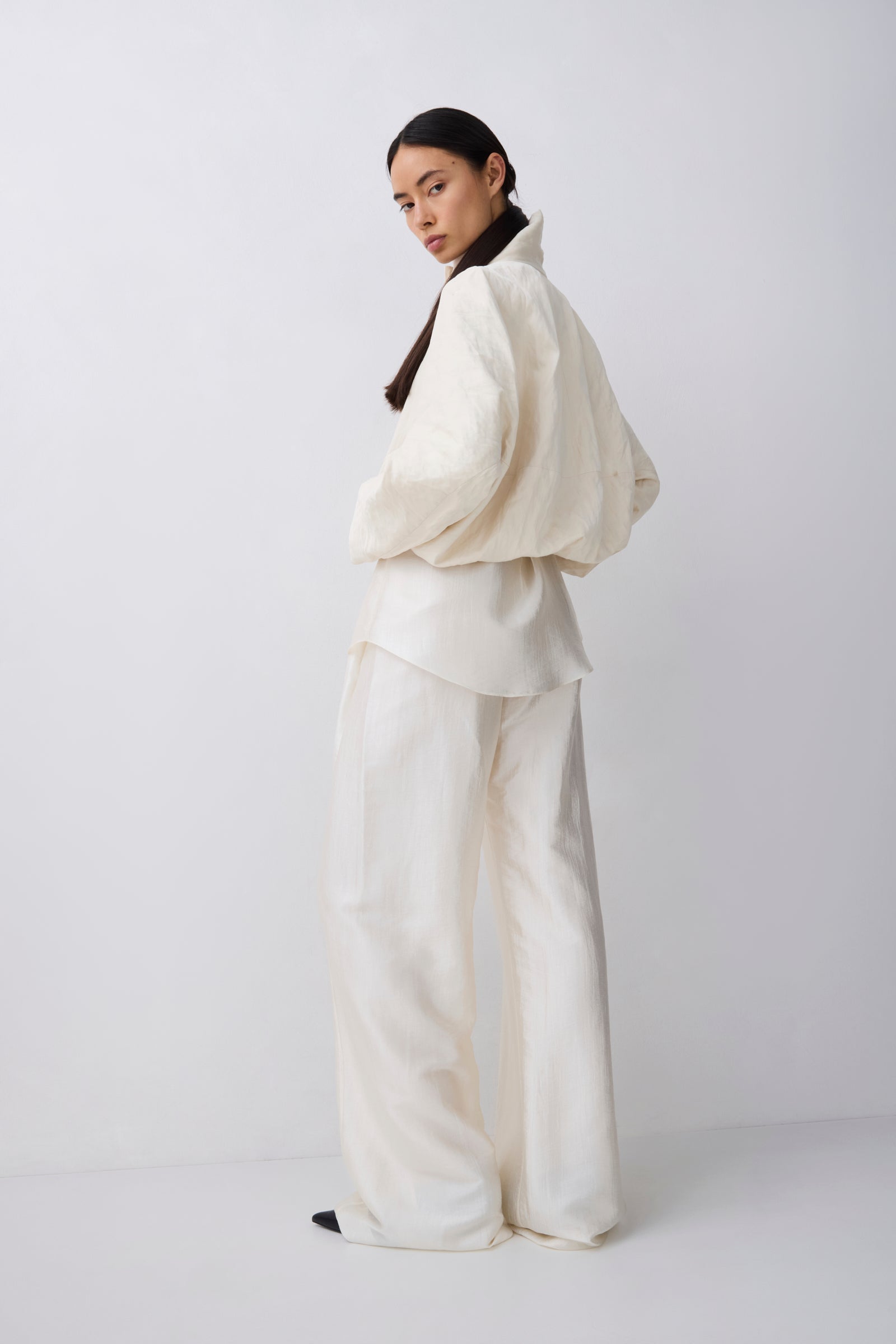 A model wearing a cream-colored two-piece outfit consisting of a voluminous long-sleeve top and matching wide-leg trousers, photographed against a plain light gray studio background.