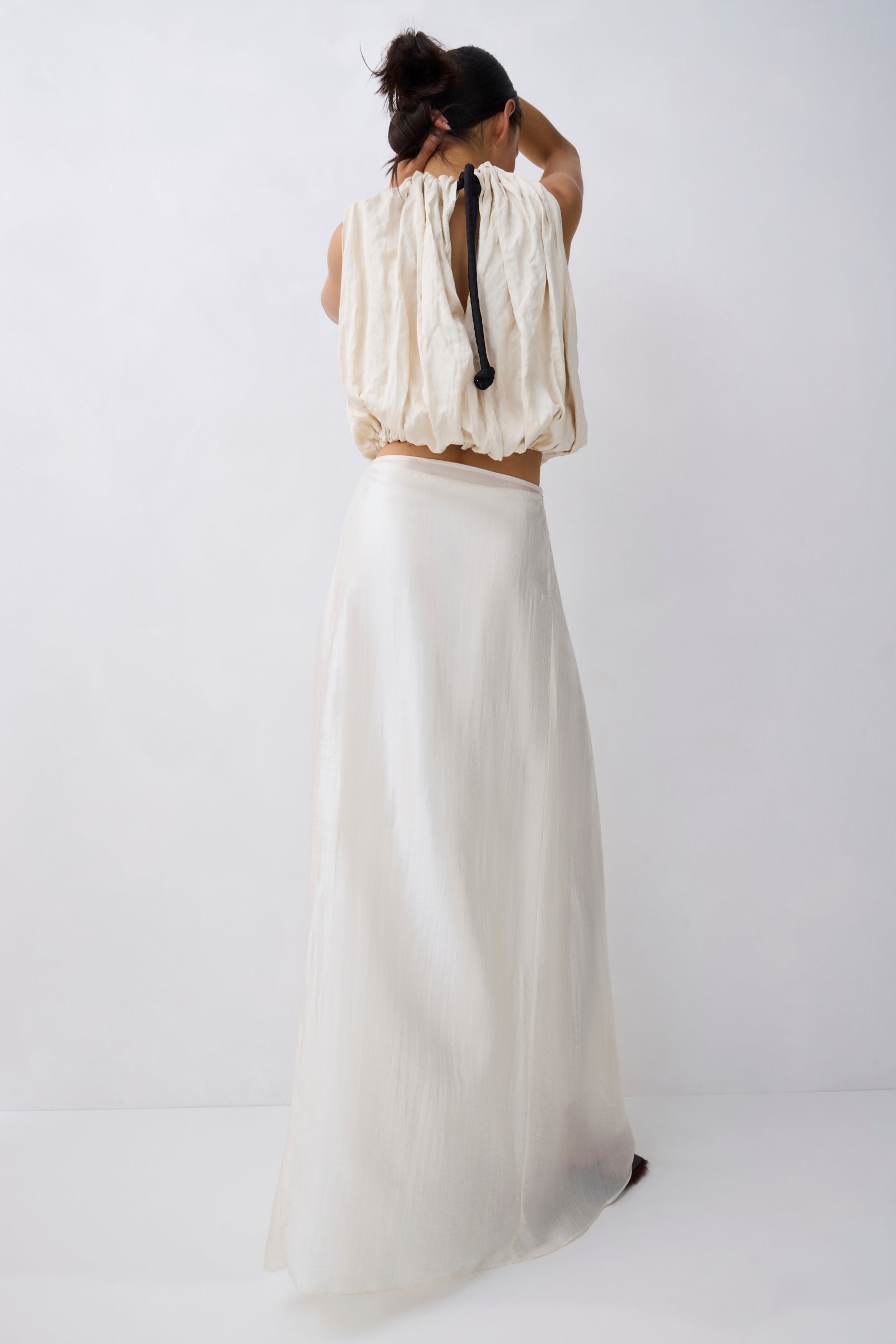 A woman shown from behind wearing a cream-colored two-piece outfit consisting of a textured, sleeveless cropped top with a tie detail and a matching long, flowing skirt, set against a plain white studio background.
