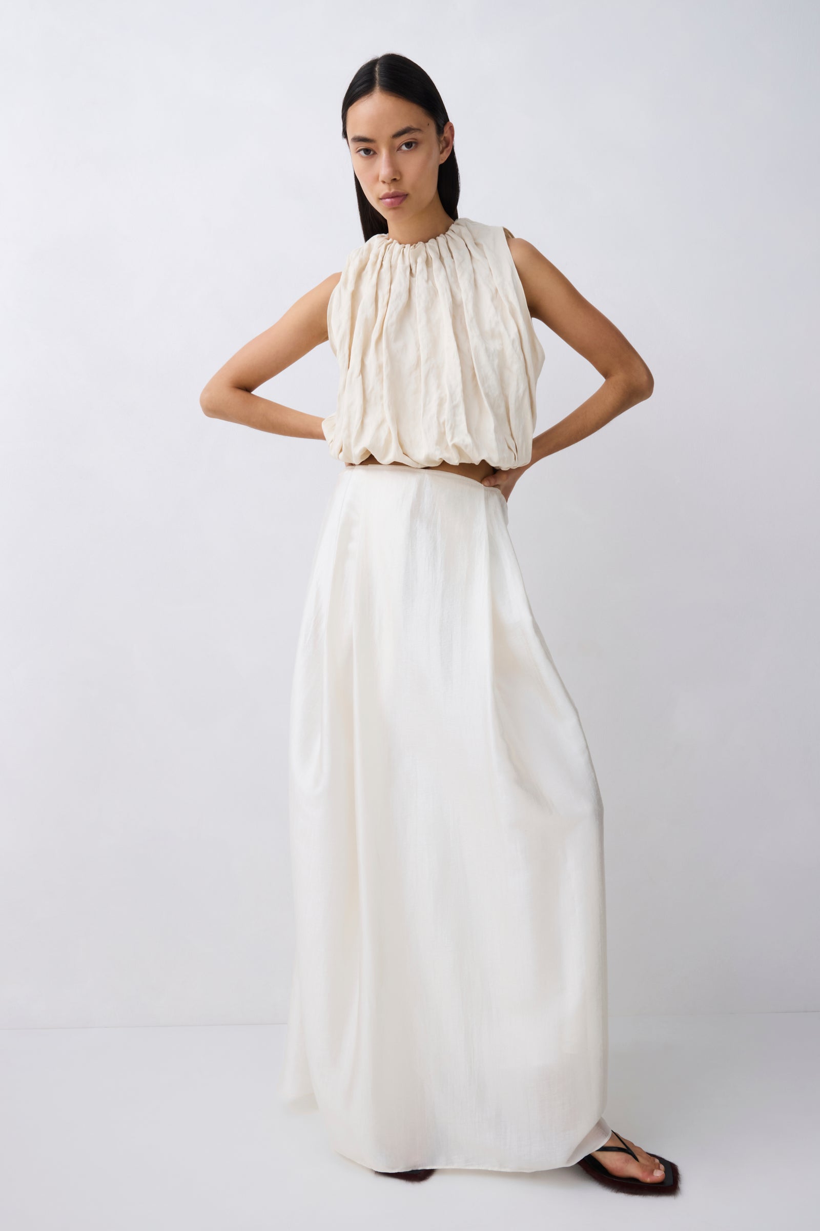 A model wearing a sleeveless, high-neck maxi dress in off-white/cream, featuring a gathered, pleated bodice and a flowing, wide-leg skirt, set against a plain light gray studio background.