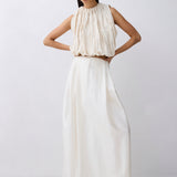 A model wearing a sleeveless, high-neck maxi dress in off-white/cream, featuring a gathered, pleated bodice and a flowing, wide-leg skirt, set against a plain light gray studio background.