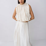 A model wearing a two-piece cream-colored outfit consisting of a sleeveless, high-neck, gathered top and a matching long skirt, photographed against a plain light gray studio background.