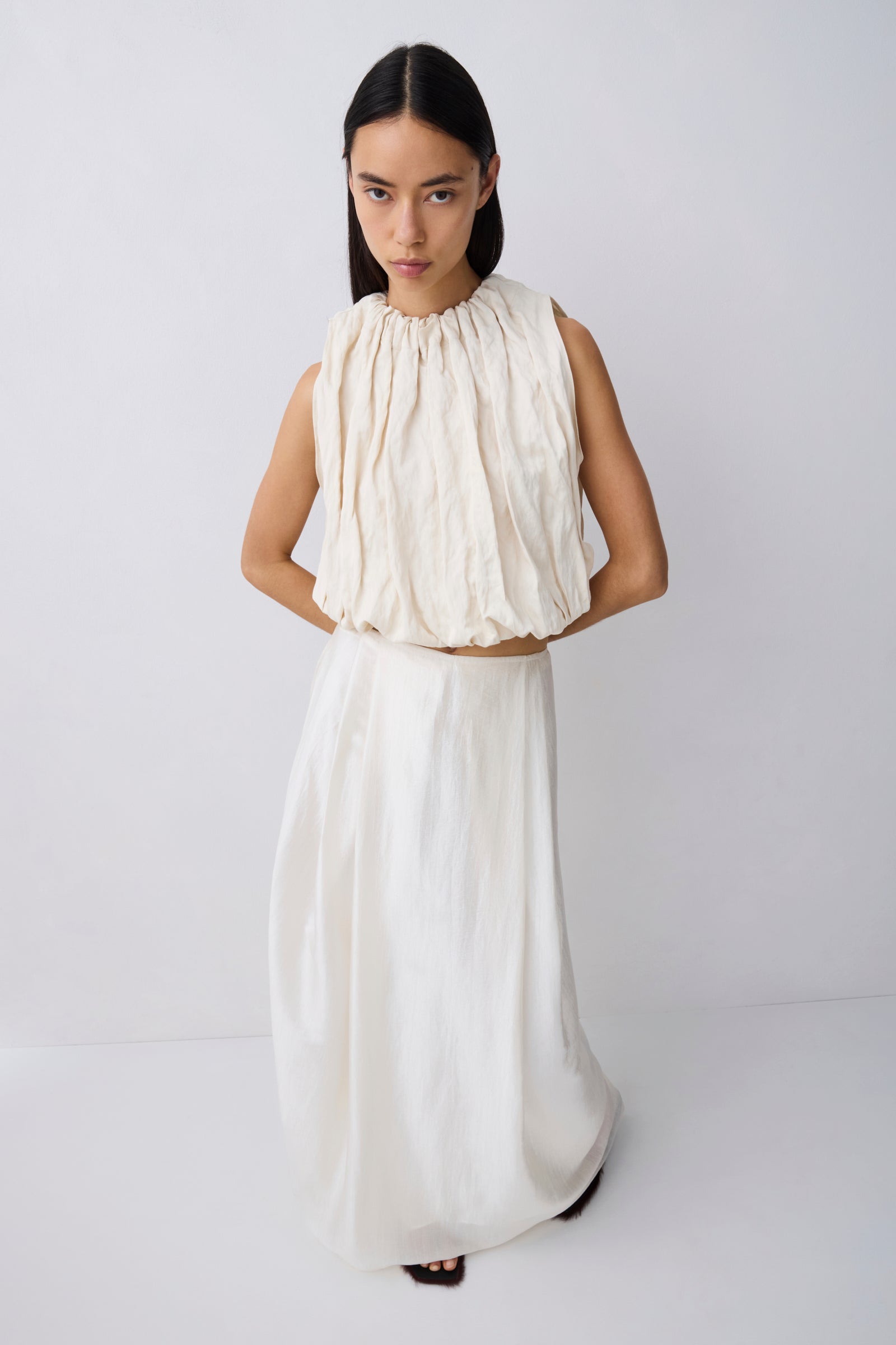 A model wearing a two-piece cream-colored outfit consisting of a sleeveless, high-neck, gathered top and a matching long skirt, photographed against a plain light gray studio background.
