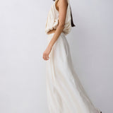A model wearing a cream-colored two-piece outfit consisting of a sleeveless draped crop top and a matching flowing maxi skirt, paired with black sandals, set against a plain light gray studio background.