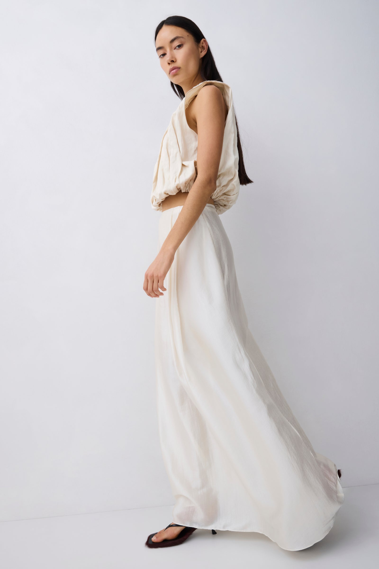 A model wearing a cream-colored two-piece outfit consisting of a sleeveless draped crop top and a matching flowing maxi skirt, paired with black sandals, set against a plain light gray studio background.