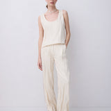A woman modeling a cream-colored linen two-piece set consisting of a sleeveless top and wide-leg trousers, standing against a plain light gray studio background.