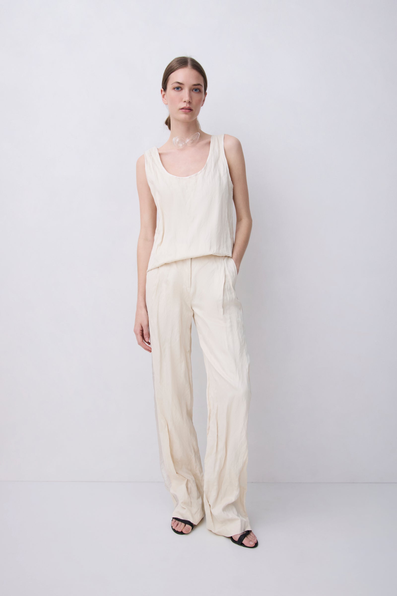 A woman modeling a cream-colored linen two-piece set consisting of a sleeveless top and wide-leg trousers, standing against a plain light gray studio background.