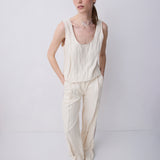 A woman modeling a cream-colored linen two-piece set consisting of a sleeveless top and wide-leg trousers, standing against a light gray studio background.