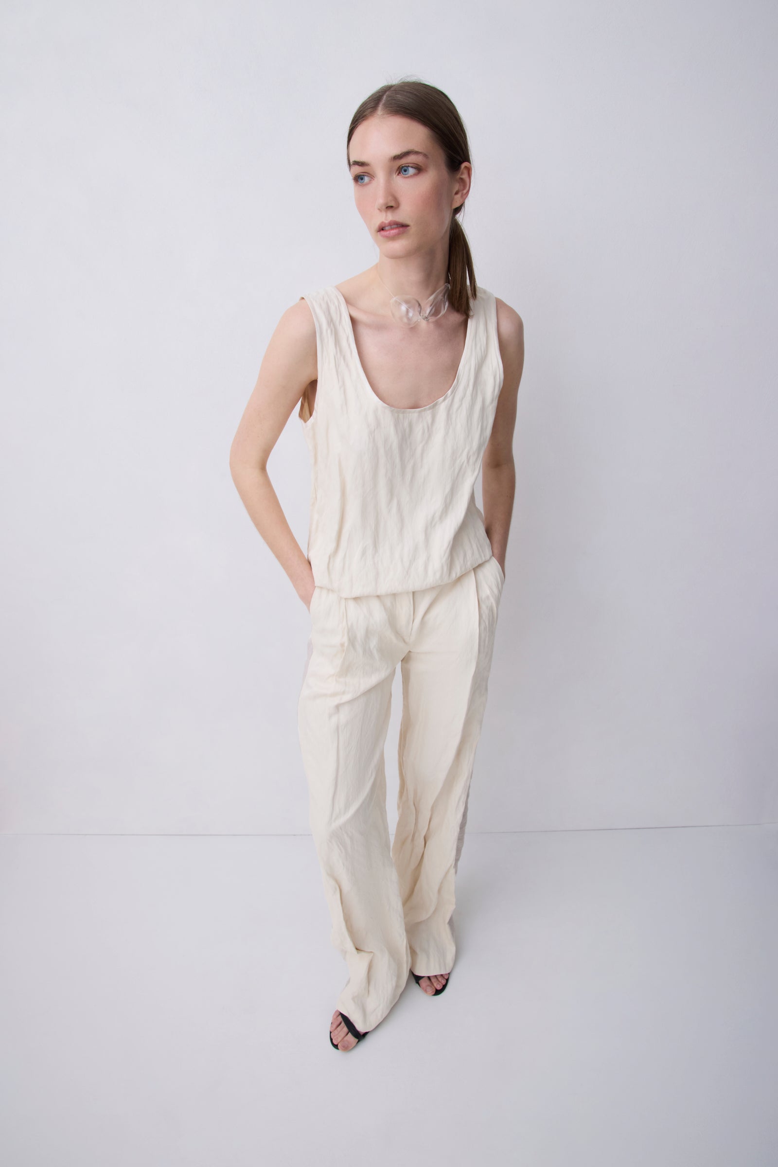 A woman modeling a cream-colored linen two-piece set consisting of a sleeveless top and wide-leg trousers, standing against a light gray studio background.