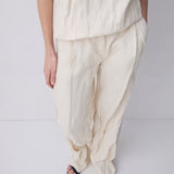 A person wearing loose, draped off-white linen pants and a matching top, paired with black strappy sandals, photographed against a light gray studio background.