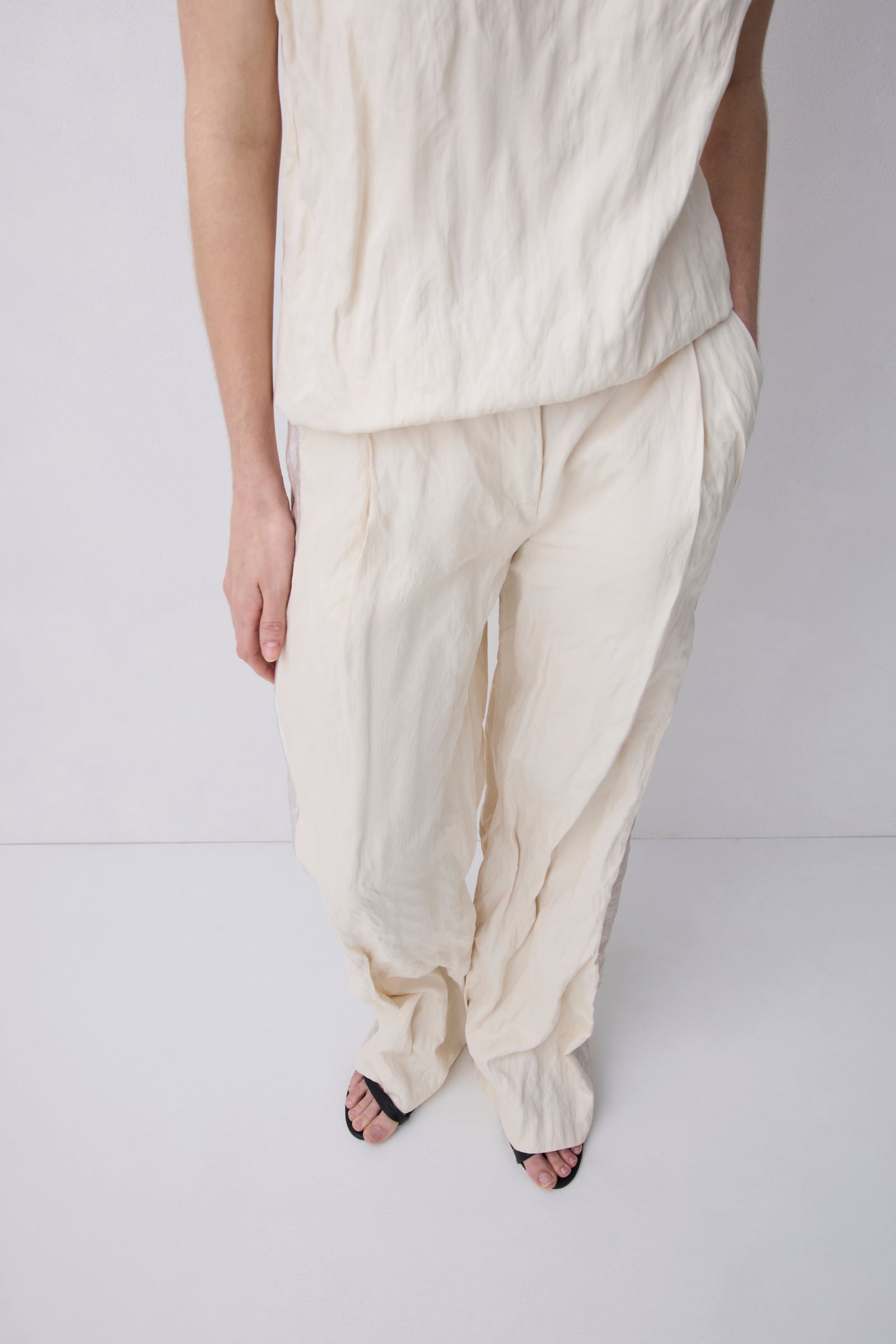 A person wearing loose, draped off-white linen pants and a matching top, paired with black strappy sandals, photographed against a light gray studio background.