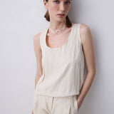A woman wearing a cream-colored sleeveless linen top and matching linen pants, standing against a light gray wall.