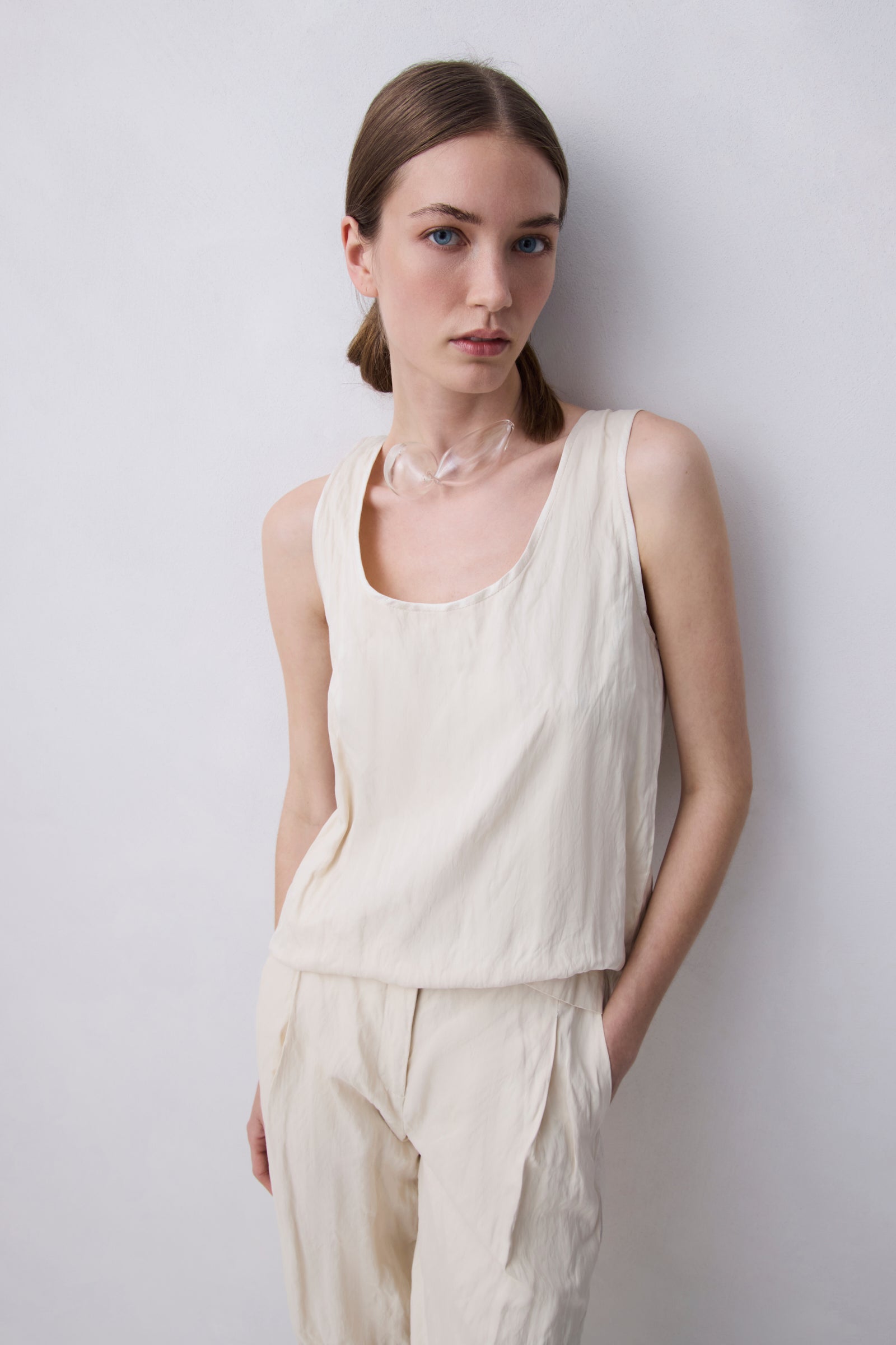 A woman wearing a cream-colored sleeveless linen top and matching linen pants, standing against a light gray wall.