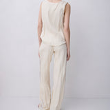 A woman stands with her back to the camera wearing a light cream-colored sleeveless top and matching wide-leg trousers against a plain light gray studio background.