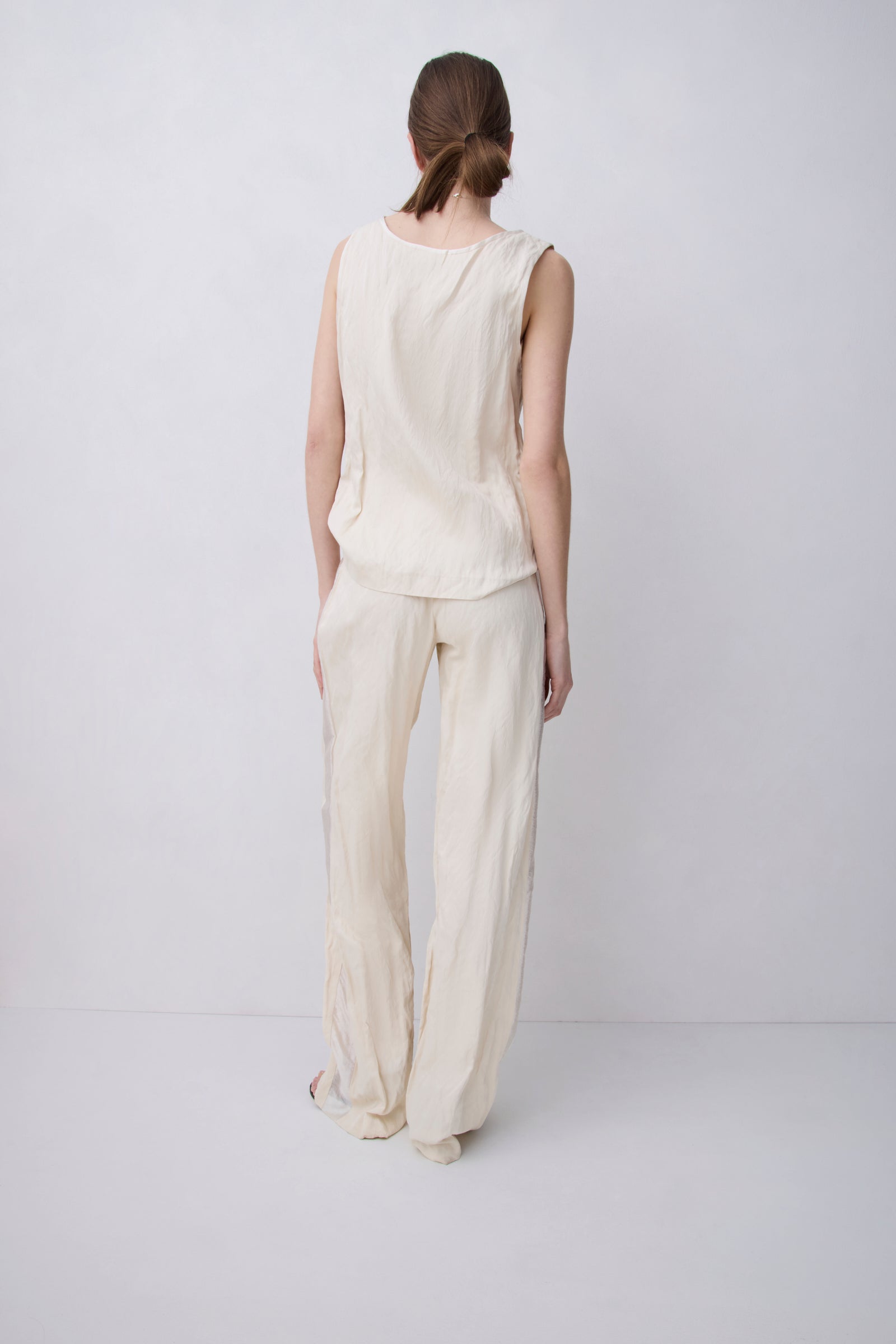 A woman stands with her back to the camera wearing a light cream-colored sleeveless top and matching wide-leg trousers against a plain light gray studio background.