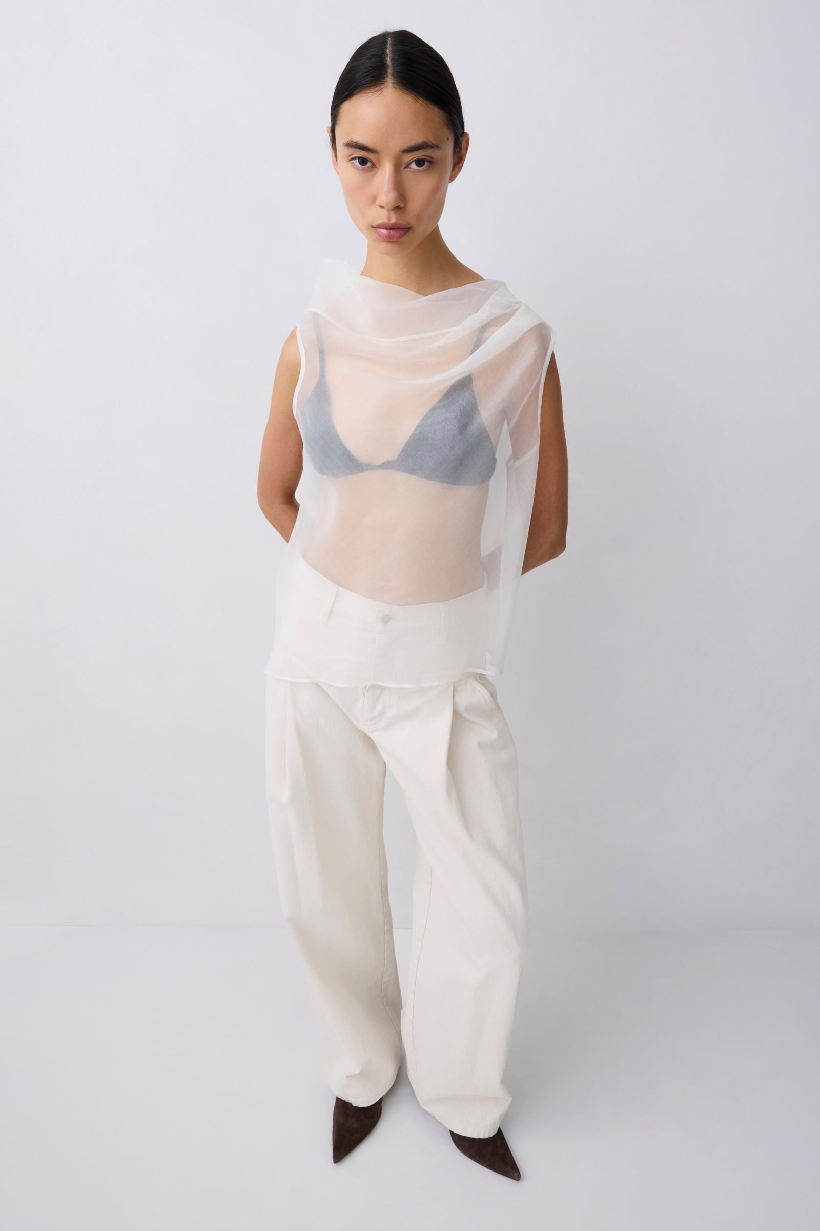 Person wearing a sheer top over a bra and white pants on a plain background