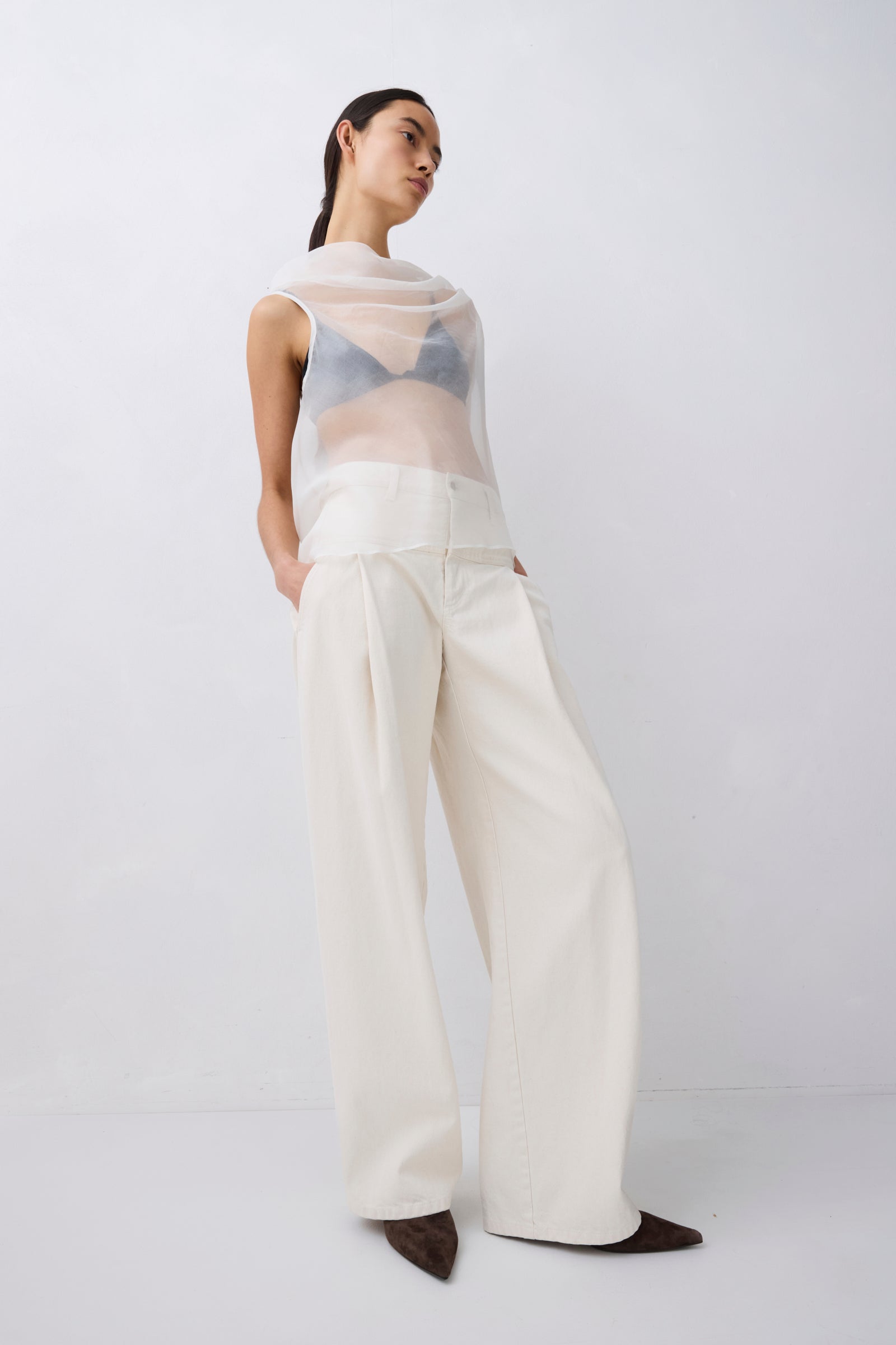 Woman wearing a sheer top and white pants on a plain background