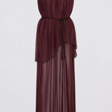 A model wearing a sleeveless, high-neck, deep burgundy maxi dress with a sheer, flowing overlay, photographed against a neutral light gray studio background.