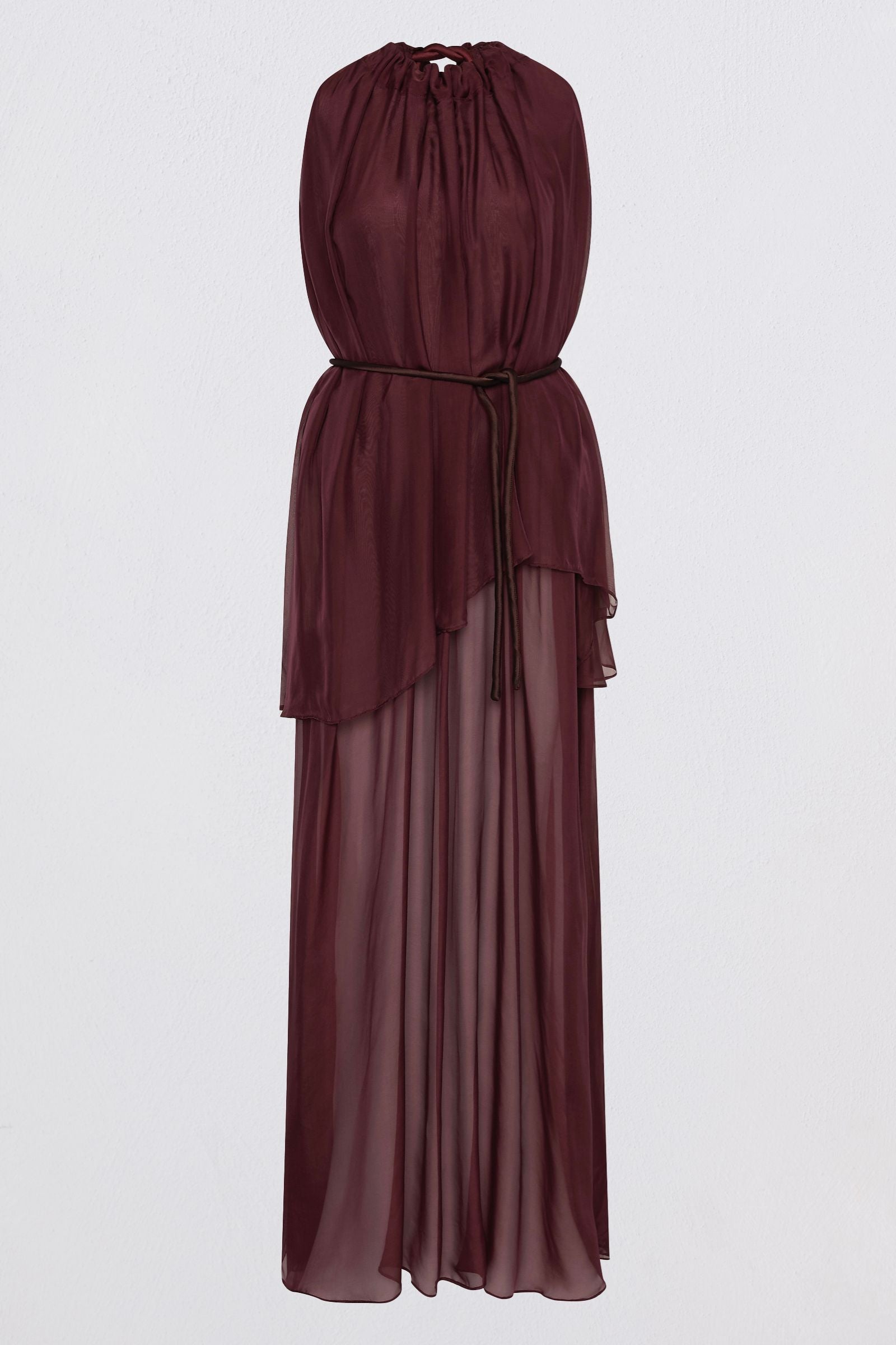 A model wearing a sleeveless, high-neck, deep burgundy maxi dress with a sheer, flowing overlay, photographed against a neutral light gray studio background.