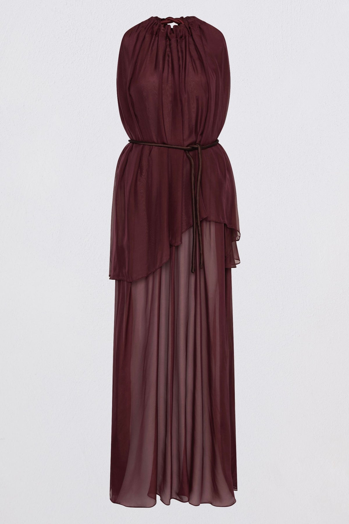 A model wearing a sleeveless, high-neck, deep burgundy maxi dress with a sheer, flowing overlay, photographed against a neutral light gray studio background.