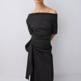 A woman modeling an off-the-shoulder black midi dress with a gathered, draped waist and asymmetrical hem, paired with black strappy sandals, against a light gray studio background.