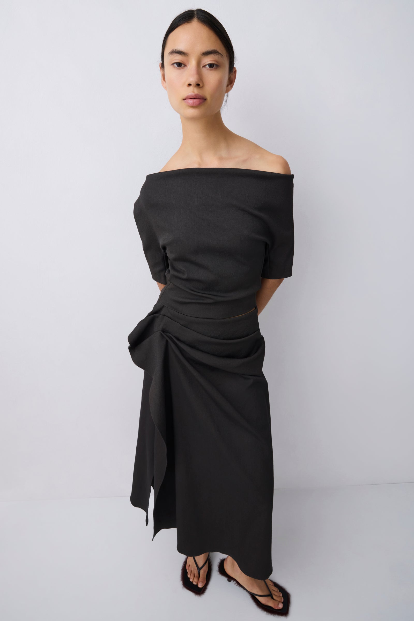 A woman modeling an off-the-shoulder black midi dress with a gathered, draped waist and asymmetrical hem, paired with black strappy sandals, against a light gray studio background.