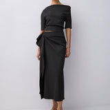 A model wearing a black off-the-shoulder midi dress with a wrap-style skirt and a matching cropped top, paired with dark brown strappy sandals, against a light gray studio background.