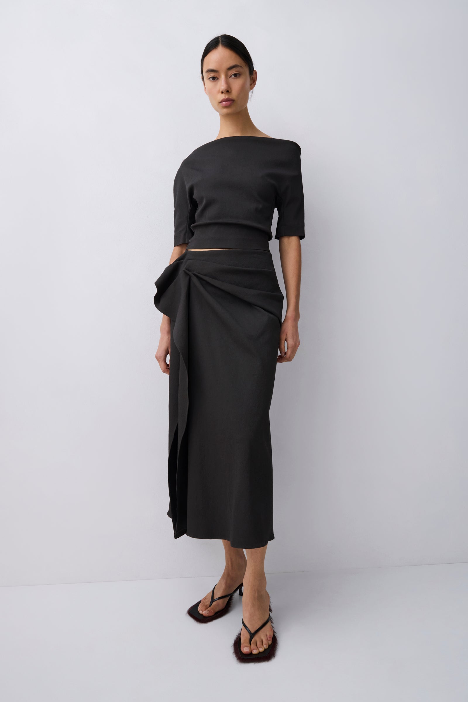 A model wearing a black off-the-shoulder midi dress with a wrap-style skirt and a matching cropped top, paired with dark brown strappy sandals, against a light gray studio background.