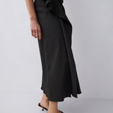 A model wearing a black wrap midi skirt with a ruffled waist, paired with black strappy mule sandals, shown from the waist down against a light gray studio background.