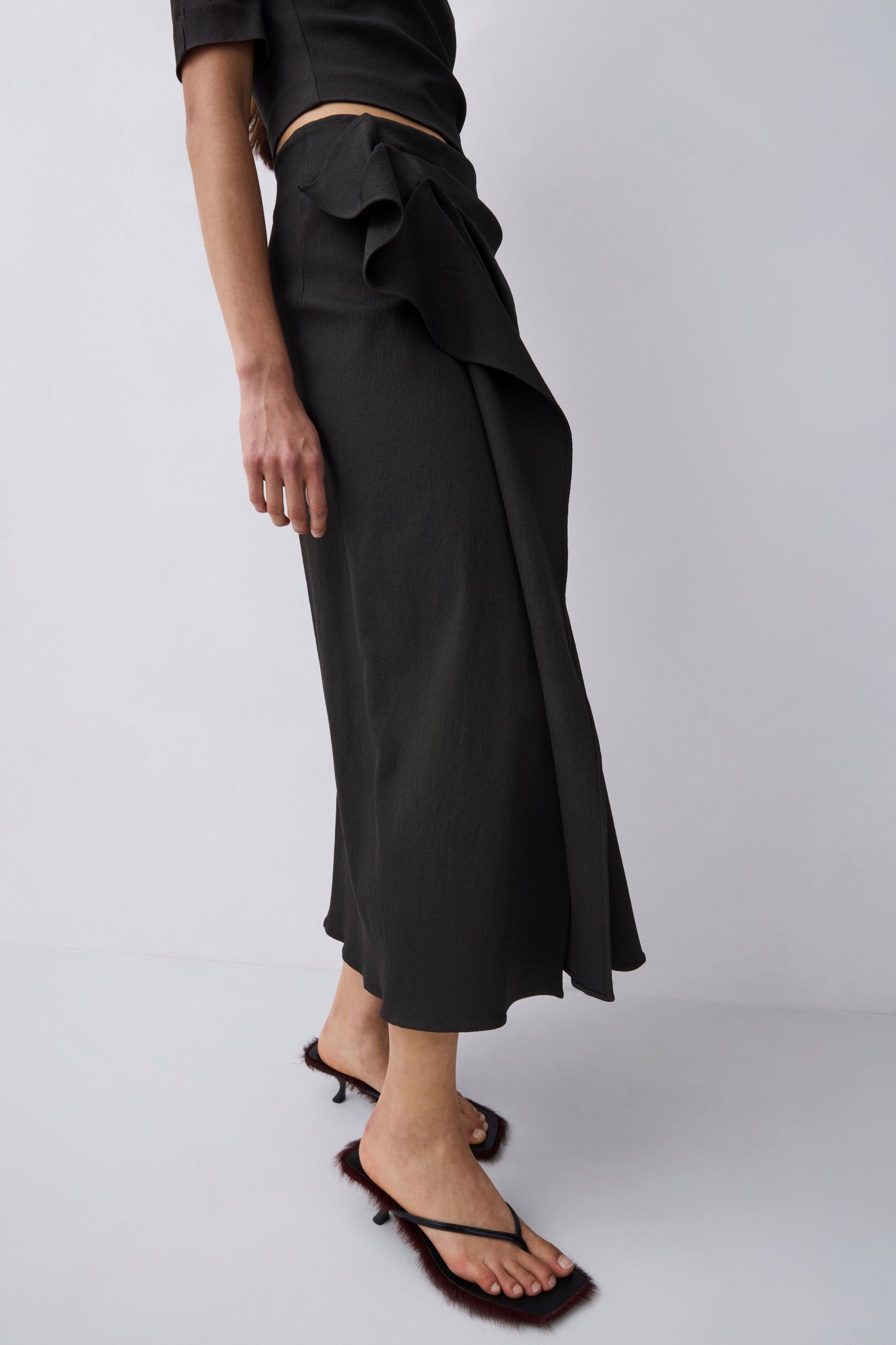 A model wearing a black wrap midi skirt with a ruffled waist, paired with black strappy mule sandals, shown from the waist down against a light gray studio background.