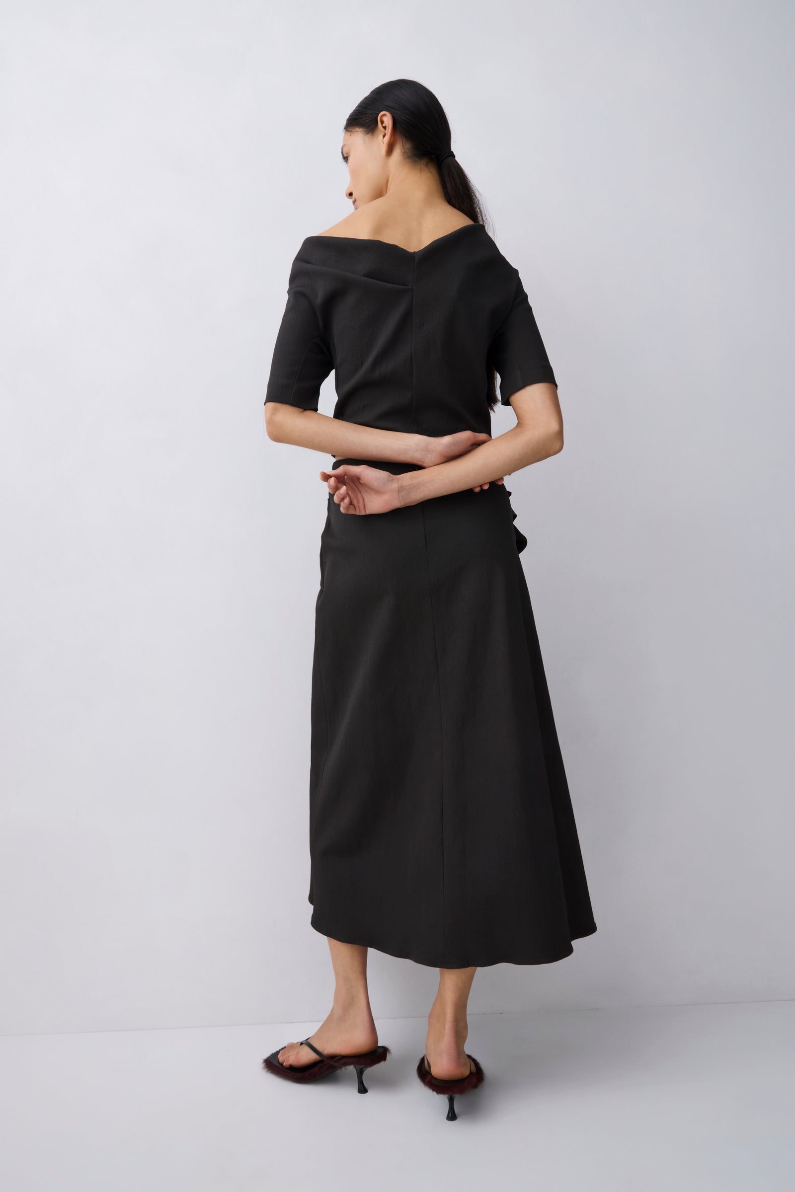 A woman modeling a black off-the-shoulder midi dress with short sleeves and a flared skirt, shown from the back against a light gray studio background.