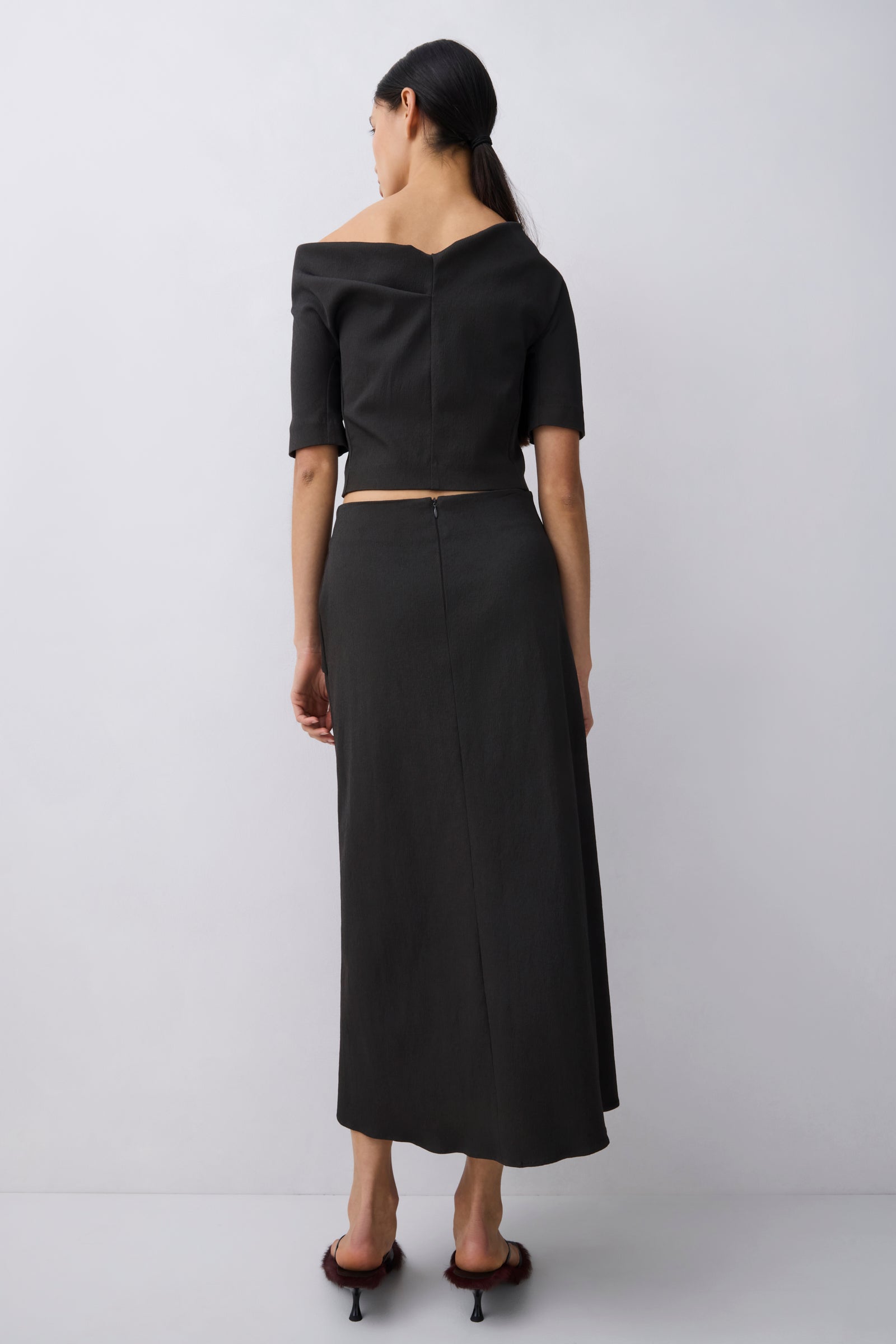 A woman stands with her back to the camera wearing a black off-the-shoulder short-sleeve crop top and a matching black midi skirt, paired with dark brown heeled sandals, against a light gray studio background.