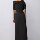 A woman stands with her back to the camera wearing a black off-the-shoulder short-sleeve crop top and a matching black midi skirt, paired with dark brown heeled sandals, against a light gray studio background.