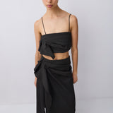 A model wearing a black two-piece outfit consisting of a strapless wrap-style crop top with a large bow detail and a matching high-waisted draped wrap skirt, paired with black flat sandals, against a plain light gray studio background.