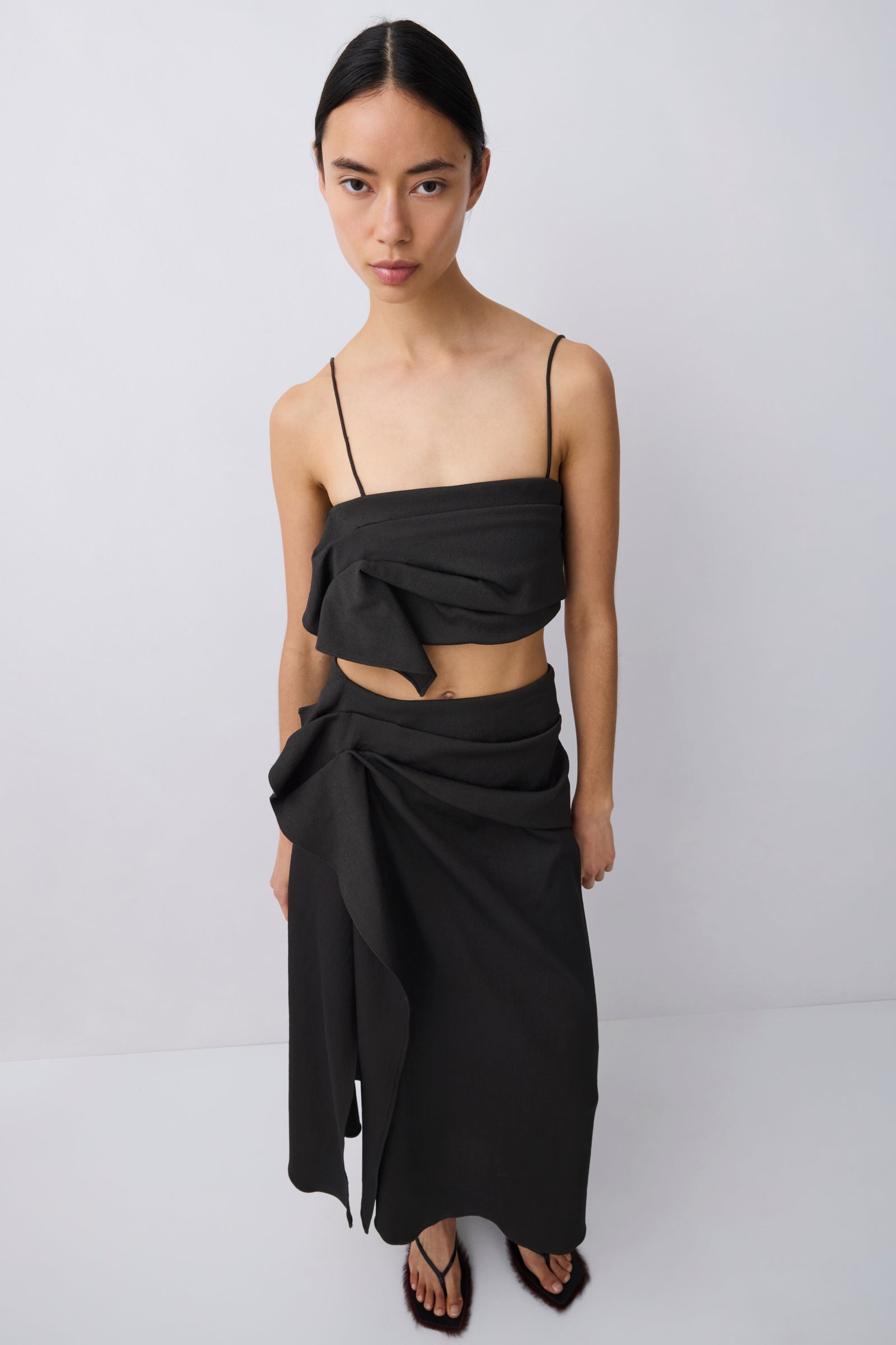 A model wearing a black two-piece outfit consisting of a strapless wrap-style crop top with a large bow detail and a matching high-waisted draped wrap skirt, paired with black flat sandals, against a plain light gray studio background.
